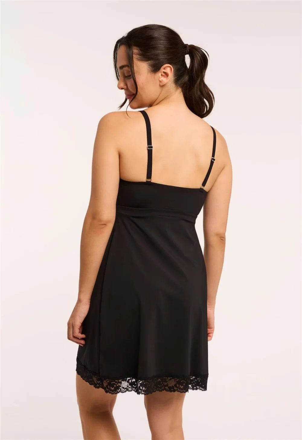 A woman shown from the back wearing a black slip-style mini dress with thin shoulder straps and a lace hem, photographed against a plain light background.
