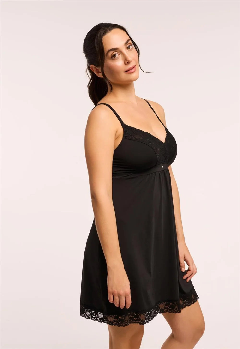 A woman modeling a black slip-style nightgown with thin straps, a lace-trimmed neckline, and a flared skirt with lace hem, photographed against a plain light background.