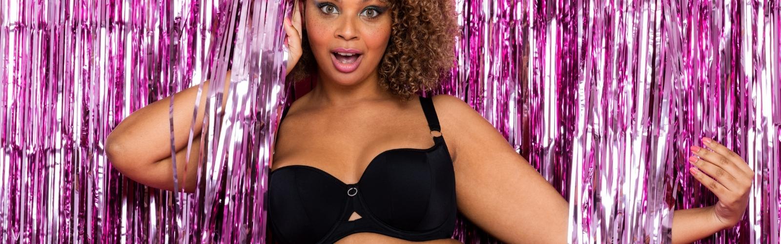 Spunky model wearing Curvy Kate Boost Me Up Padded Balcony Bra in black (CK0271), striking a confident pose against a fun pink party-themed background – featured on Bravo Bra Boutique.